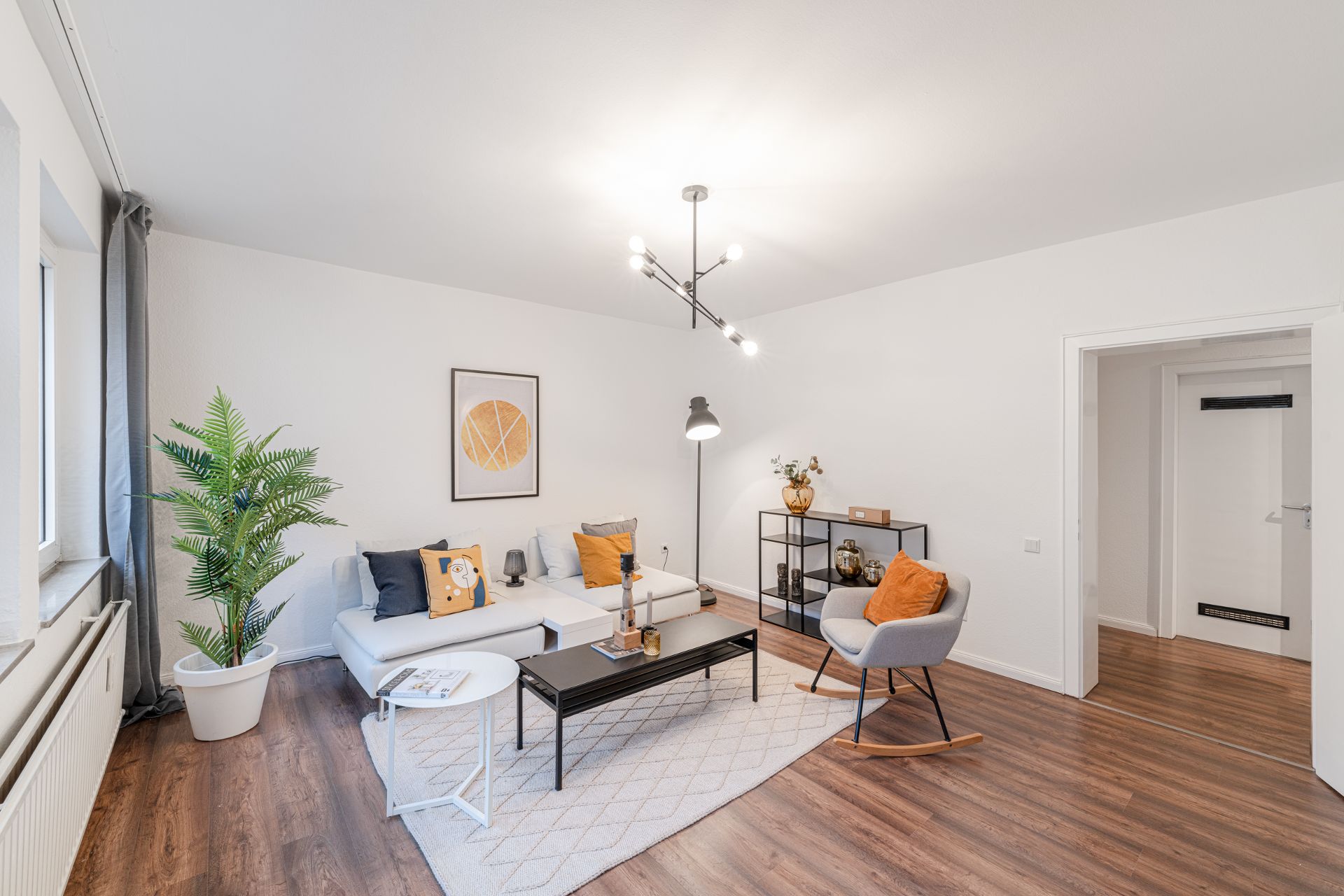 Helles Wohnzimmer mit Sofa, Sessel, Couchtisch, Regal, Pflanze und Holzboden.