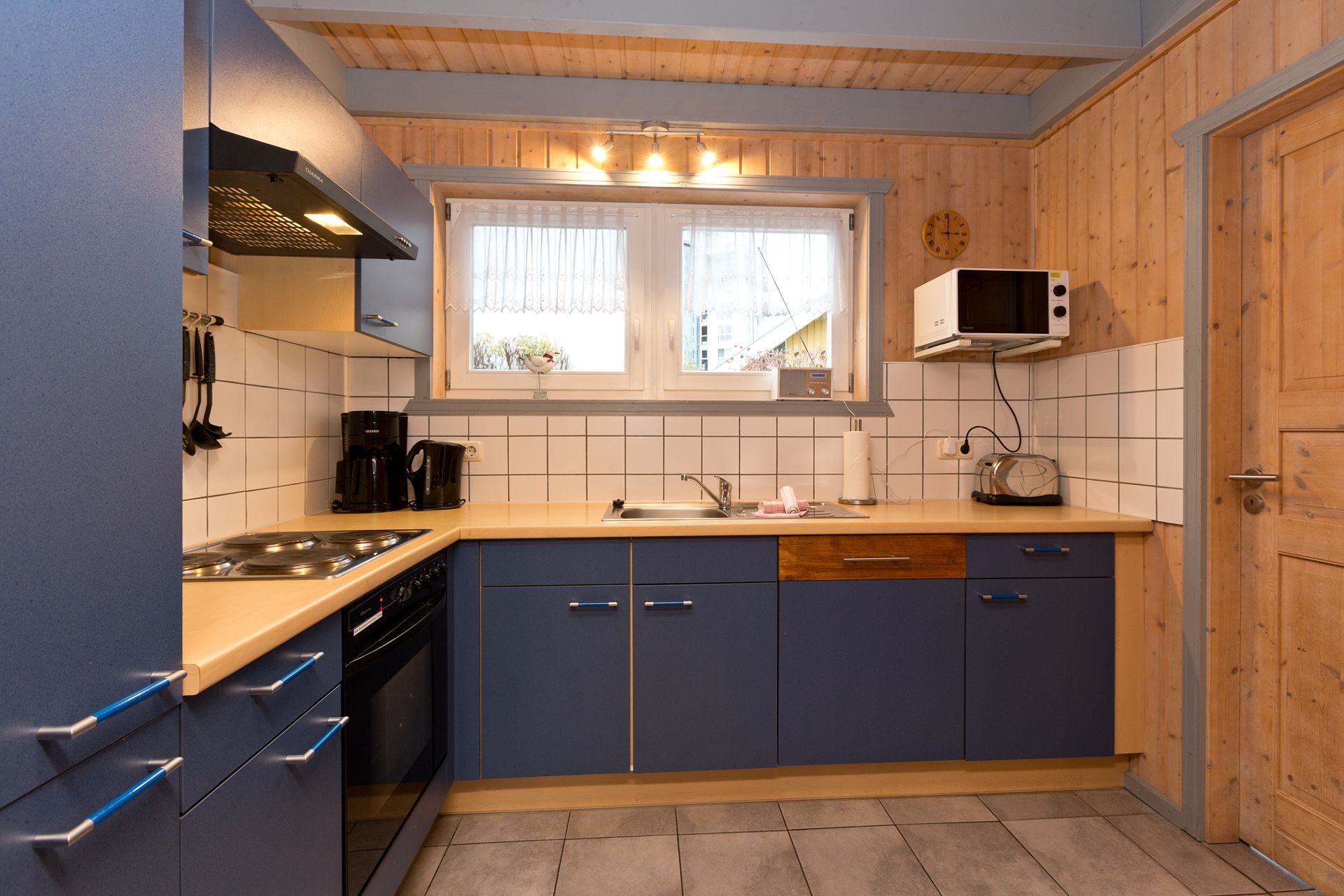 Küche mit blauen Schränken, Herd, Spüle, Kaffeemaschine, Mikrowelle und Toaster unter einem Fenster.