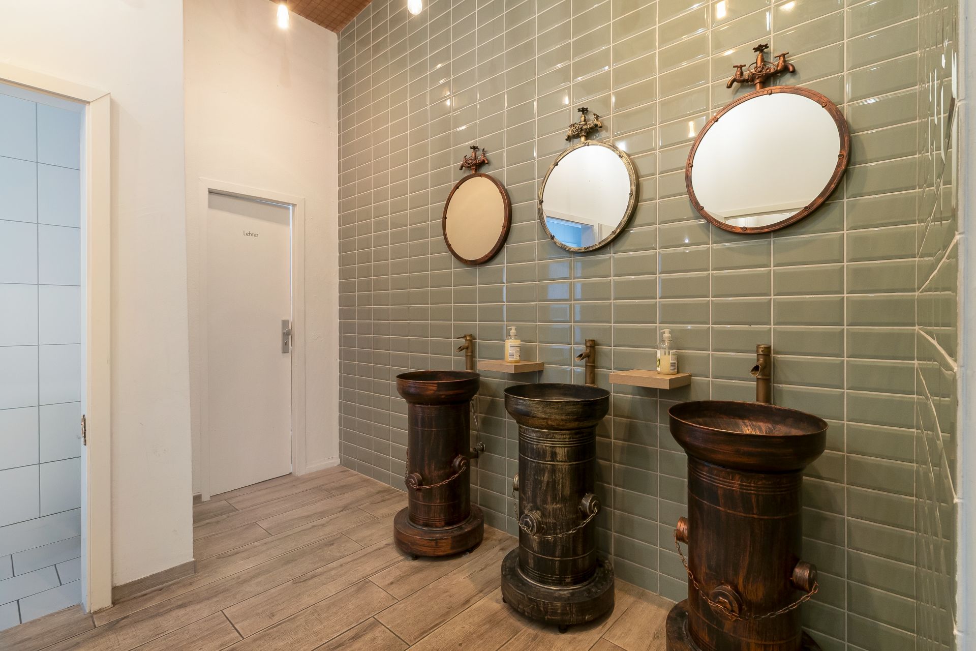 Drei runde Spiegel und drei Waschbecken vor grüner Fliesenwand in einem Badezimmer.