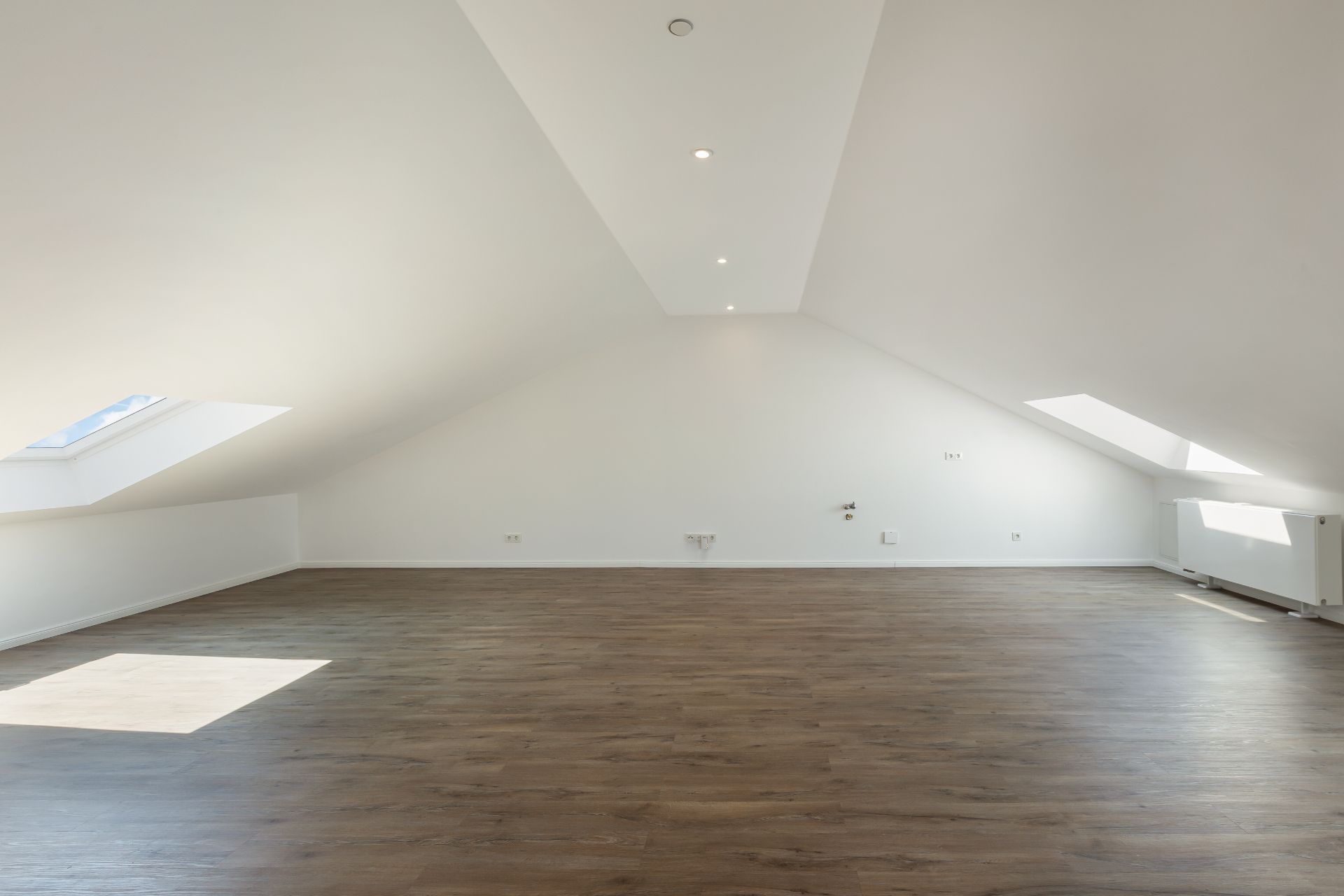 Heller Dachbodenraum mit schrägen weißen Wänden, Holzboden und zwei Dachfenstern.