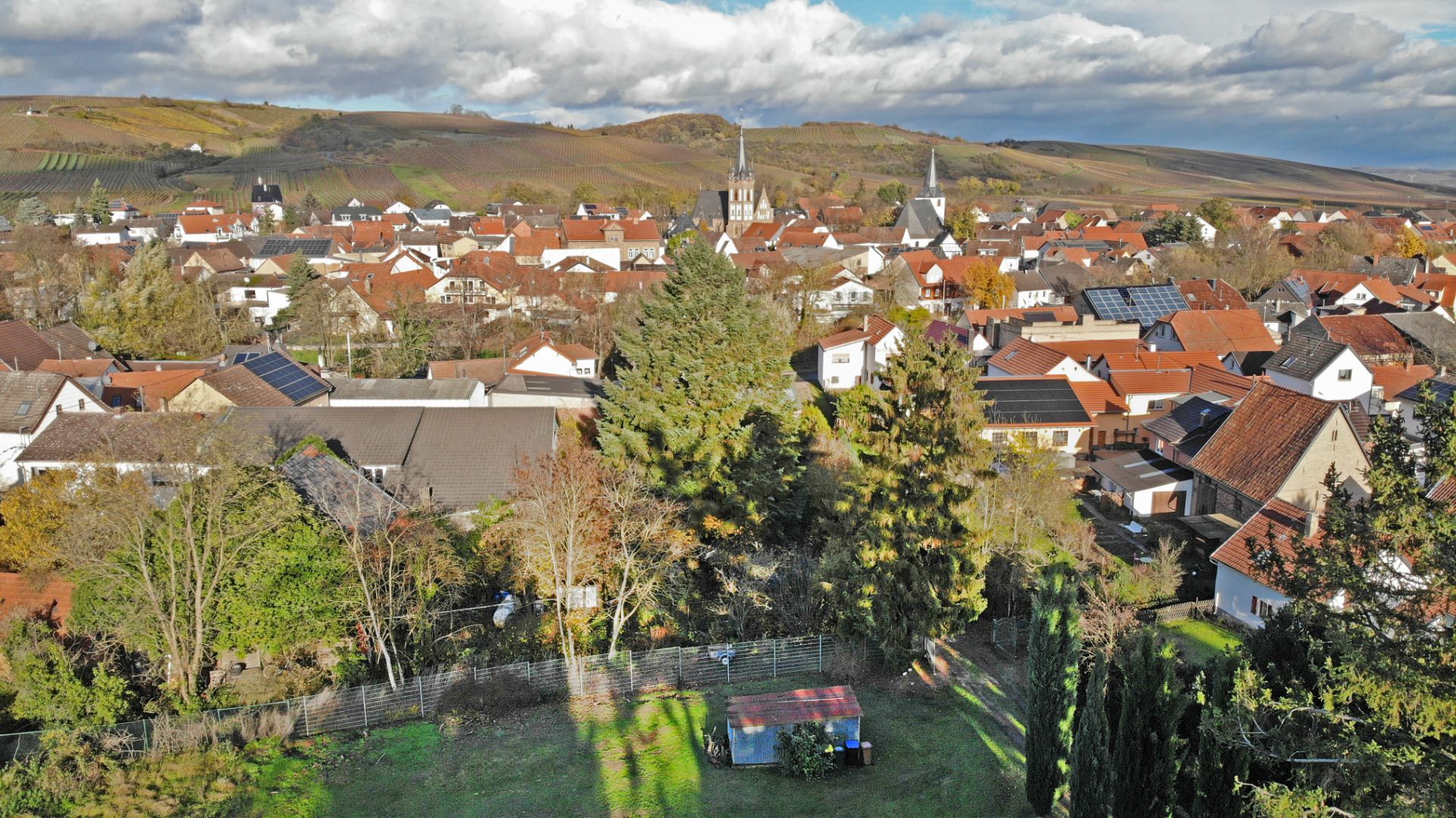 Blick auf ein Wohngebiet mit roten Dächern, Bäumen und Hügeln im Hintergrund.