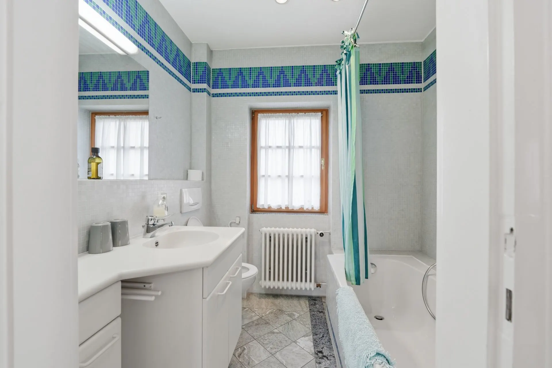 Helles Badezimmer mit Waschbecken, Spiegel, Badewanne, Fenster und blauem Fliesenband.