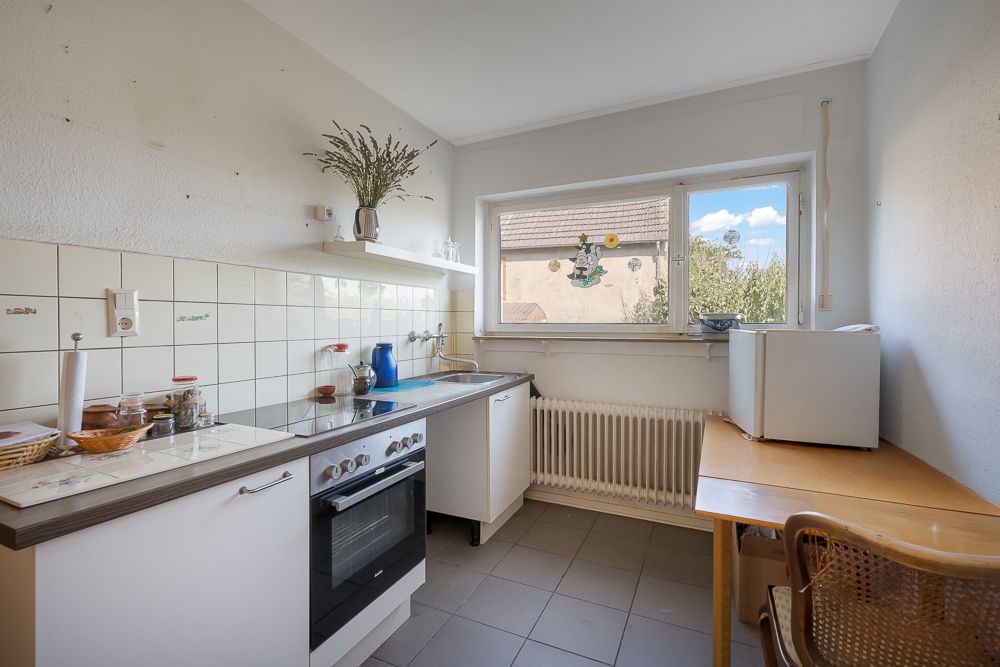 Helle Küche mit Herd, Spüle, Kühlschrank, Tisch und Stuhl, und großem Fenster zur Außenseite.