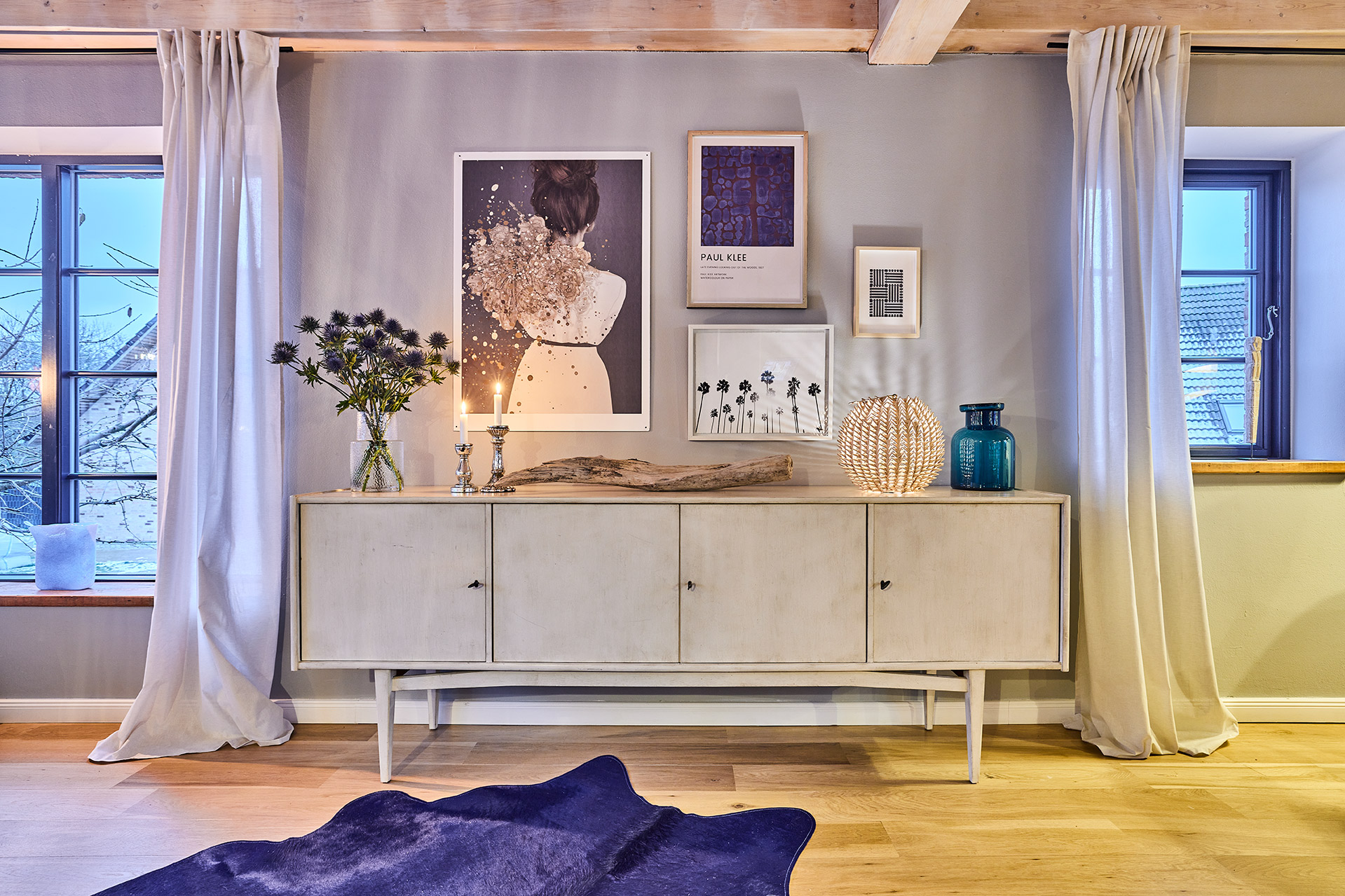 Wohnzimmer mit Sideboard, Blumen, Kerzen, Dekorationen und Wandbildern, zwischen zwei Fenstern mit Vorhängen.
