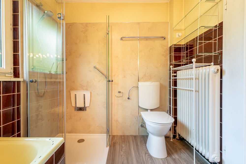 Helles Badezimmer mit Dusche, Toilette, Heizkörper und Regalen an der Wand.