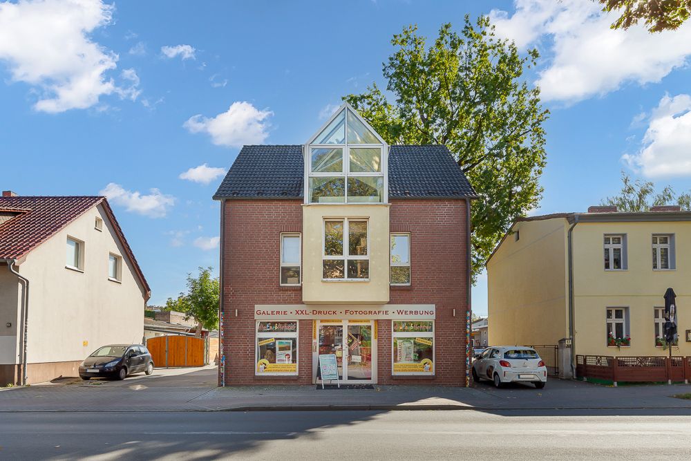 Zweistöckiges Haus mit roter Fassade und großem Fenster im Dachgeschoss, Geschäfte im Erdgeschoss.