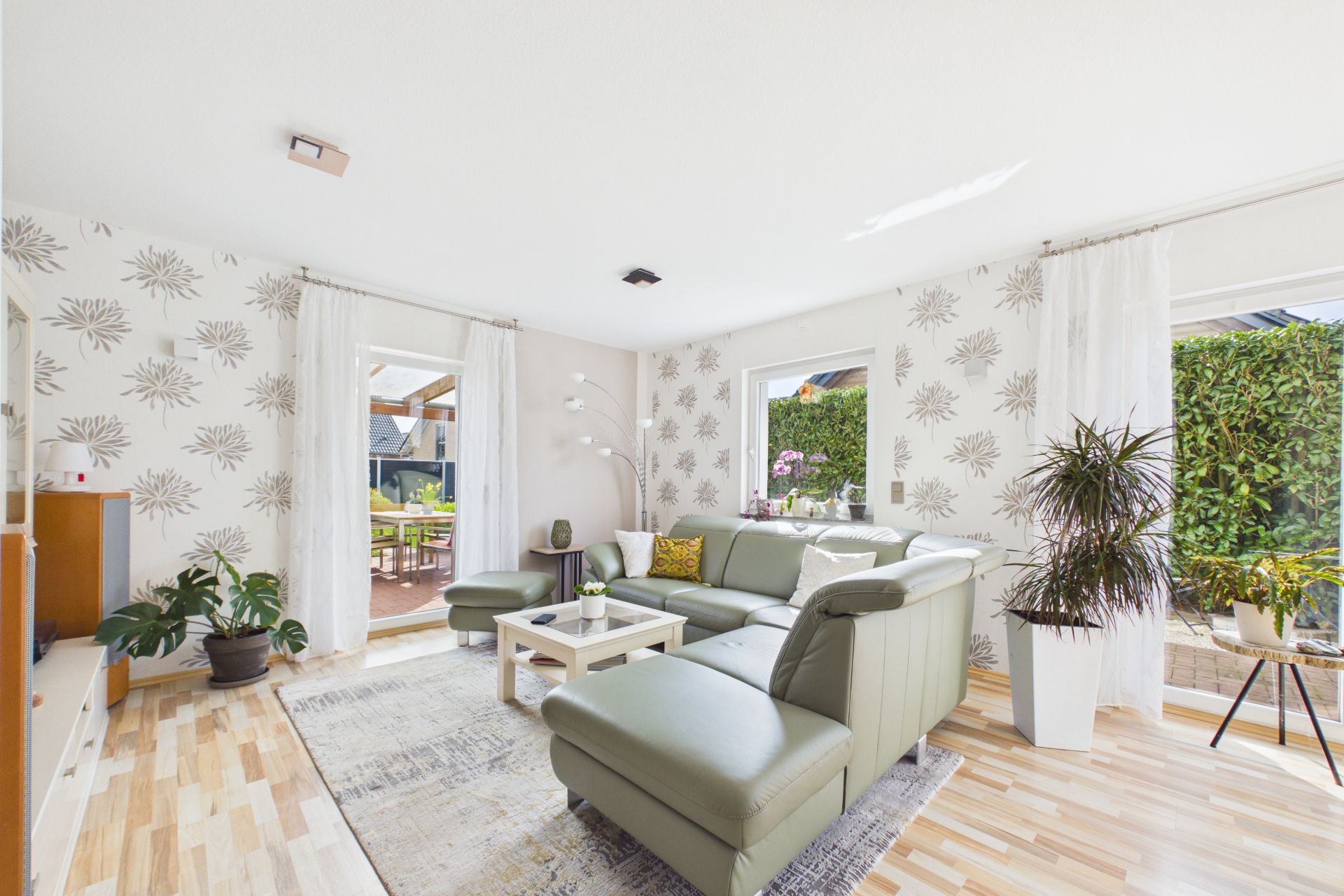 Helles Wohnzimmer mit grüner Eckcouch, Pflanzen und großem Fenster zum Garten.