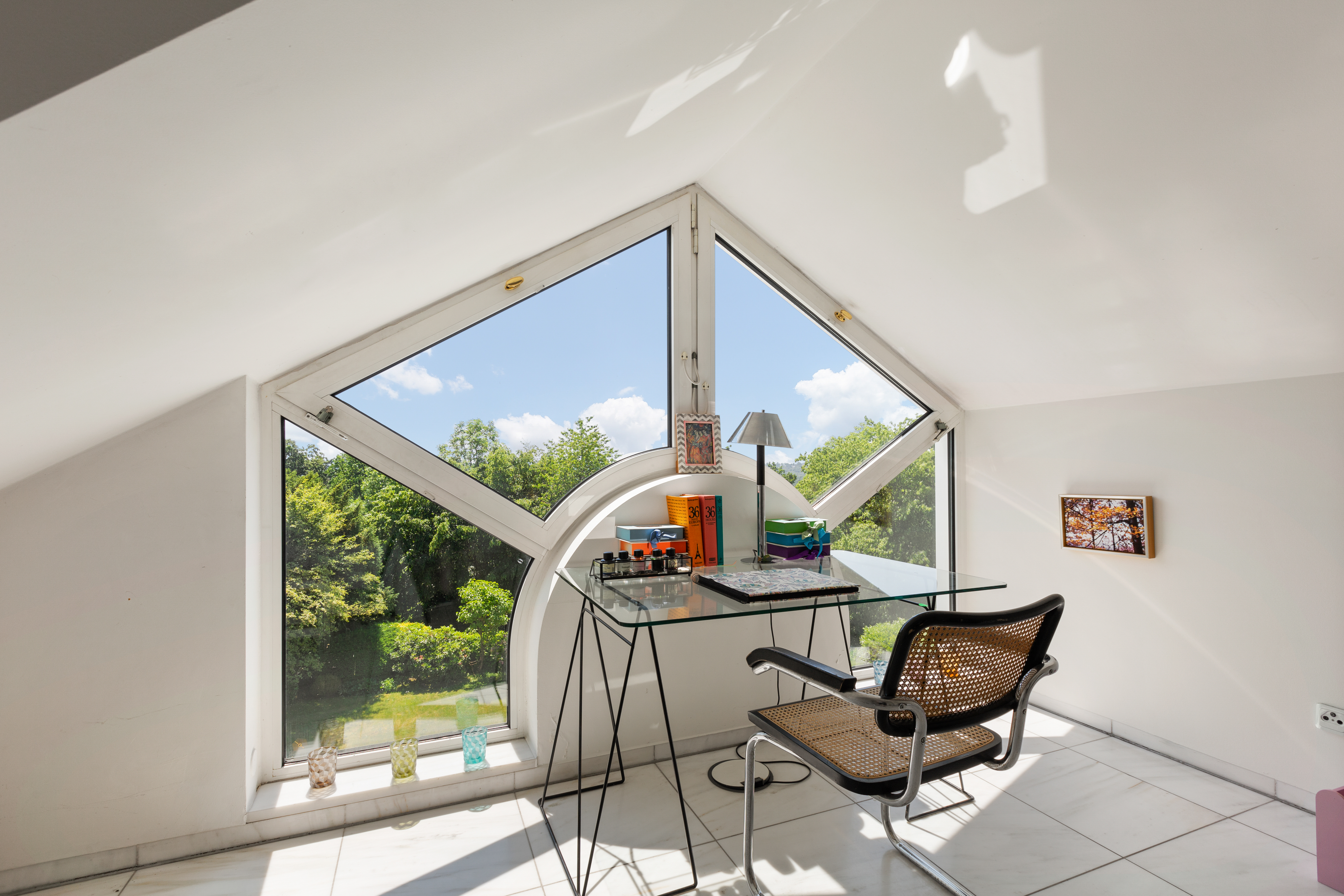 Heller Arbeitsbereich mit Glas-Schreibtisch vor großen, geometrischen Fenstern mit Blick ins Grüne.