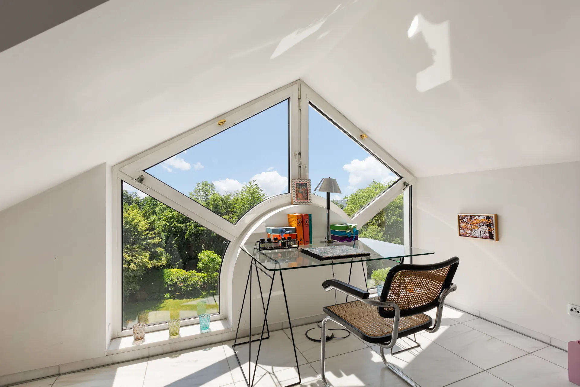 Heller Arbeitsbereich mit Glas-Schreibtisch vor großen, geometrischen Fenstern mit Blick ins Grüne.