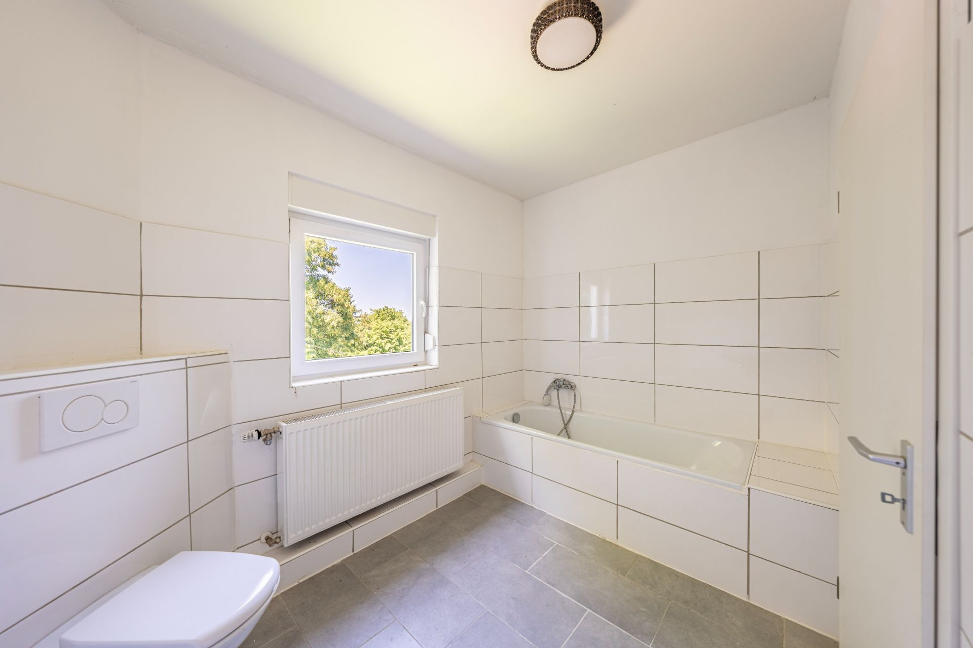 Helles Badezimmer mit Badewanne, Toilette, Fenster und weißen Wandfliesen.