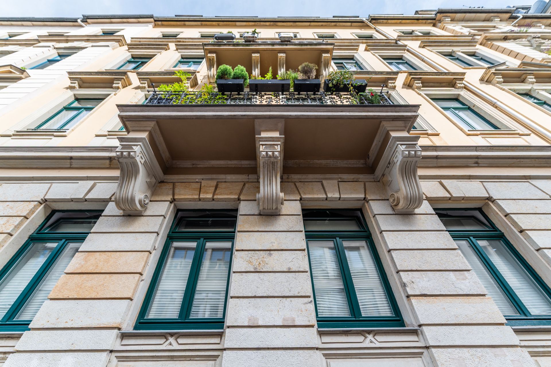 Blick von unten auf eine Hausfassade mit grün gerahmten Fenstern und Balkon mit Pflanzen.