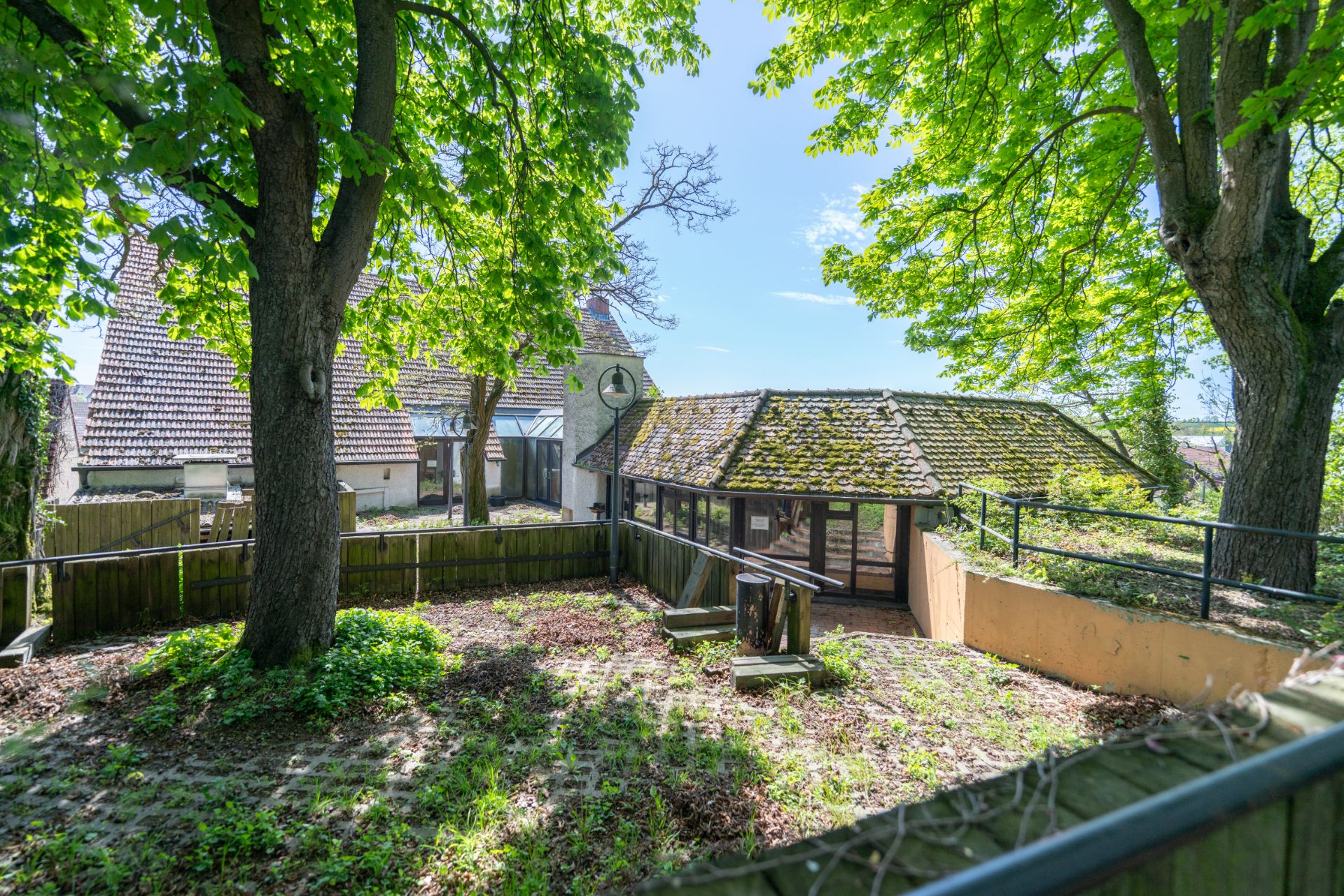 Garten mit großen Bäumen, Holzzaun und Blick auf ein Haus mit Ziegeldach.