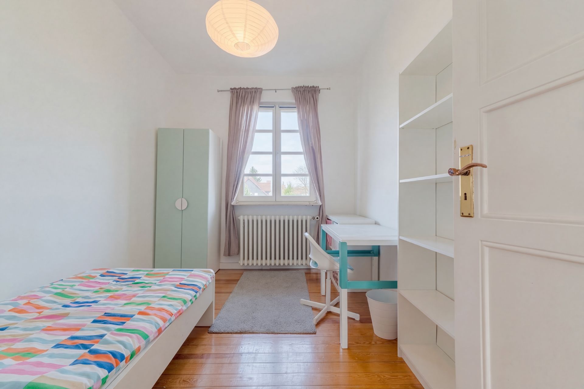 Helles Zimmer mit Einzelbett, Schreibtisch, Regal und Fenster mit Vorhängen.