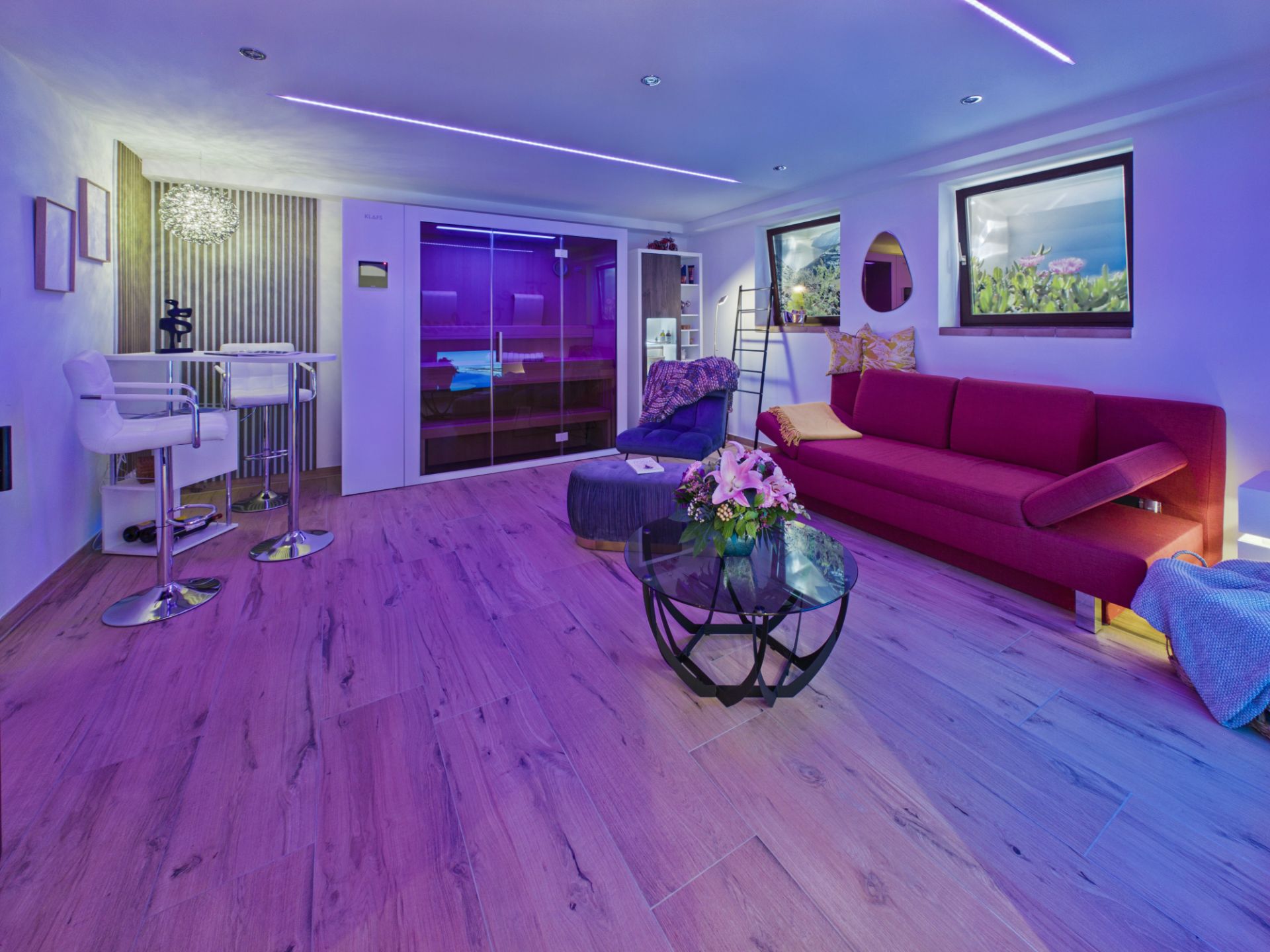 Modernes Wohnzimmer mit lila Beleuchtung, rotem Sofa und Glastisch, Barhockern und großen Fenstern.
