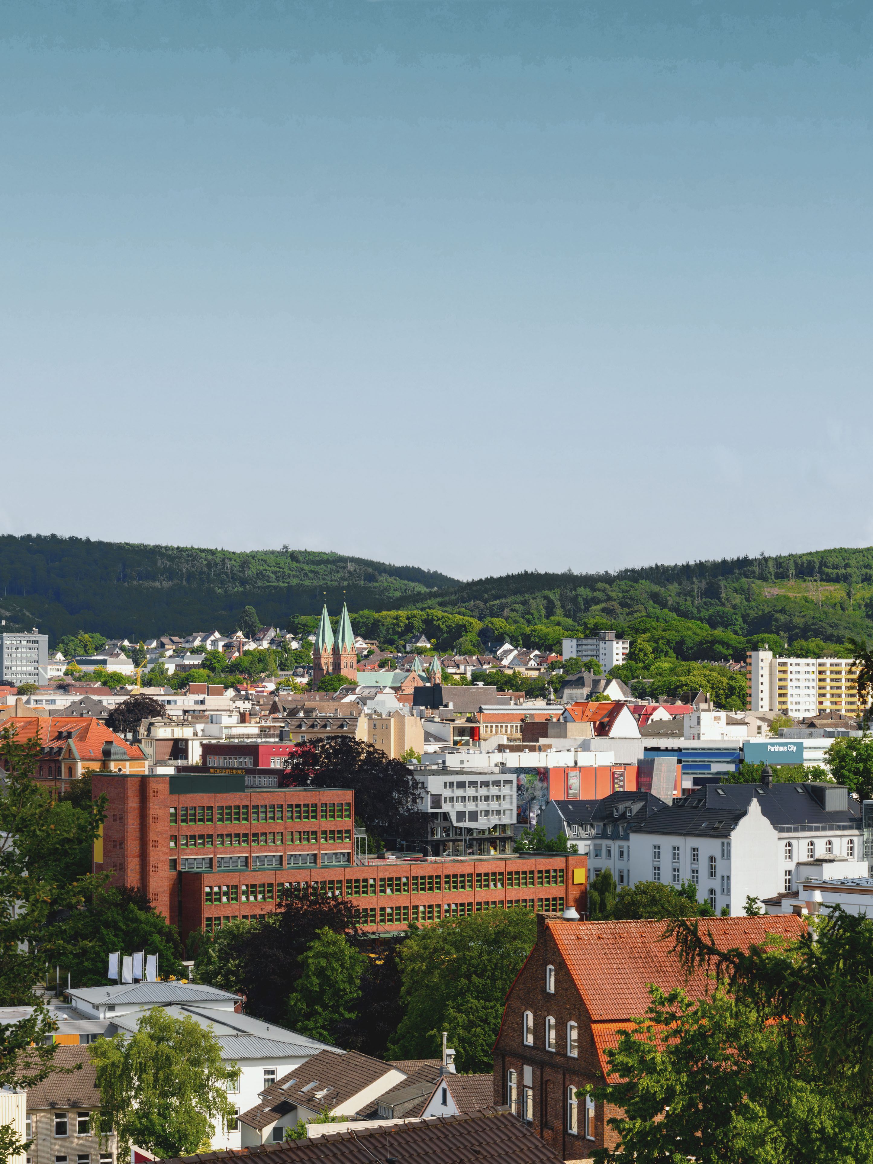 Stadtpanorama mit Wohngebäuden, roten Dächern und grünen Hügeln im Hintergrund.