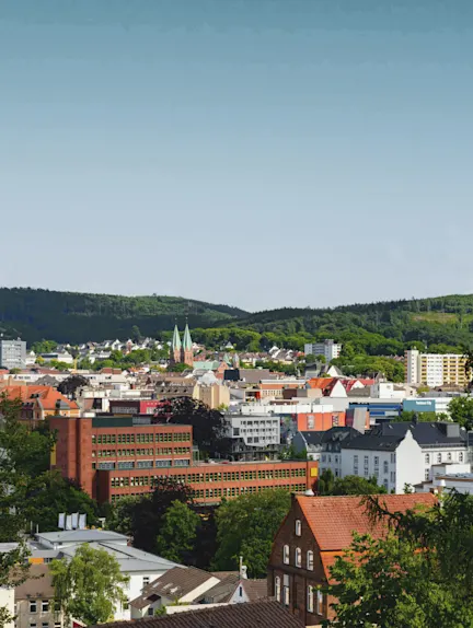 Stadtpanorama mit Wohngebäuden, roten Dächern und grünen Hügeln im Hintergrund.