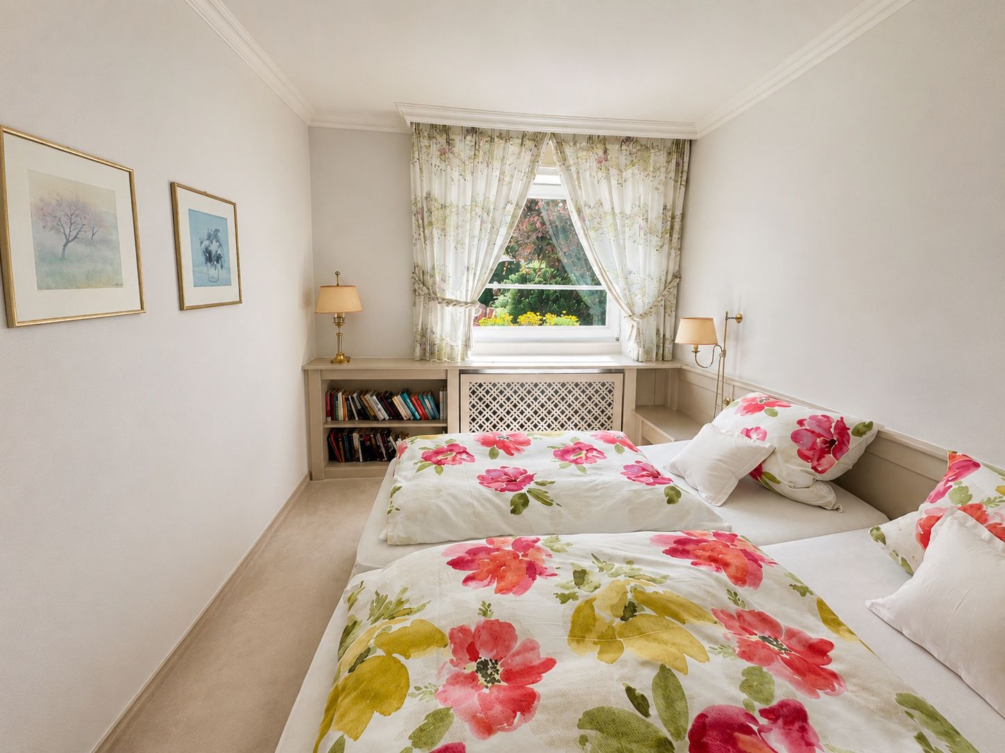 Helles Schlafzimmer mit zwei Betten, Blümchenbettwäsche, Bücherregal und Fenster mit Vorhängen.