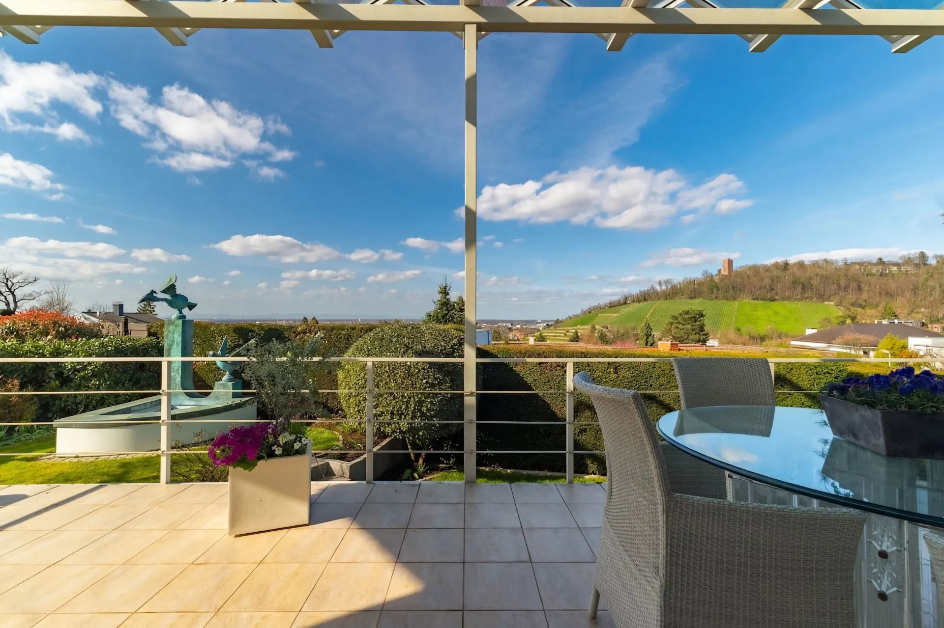 Balkon mit Glas-Esstisch, Stühlen und Ausblick auf einen Garten und grüne Hügel bei Sonnenschein.