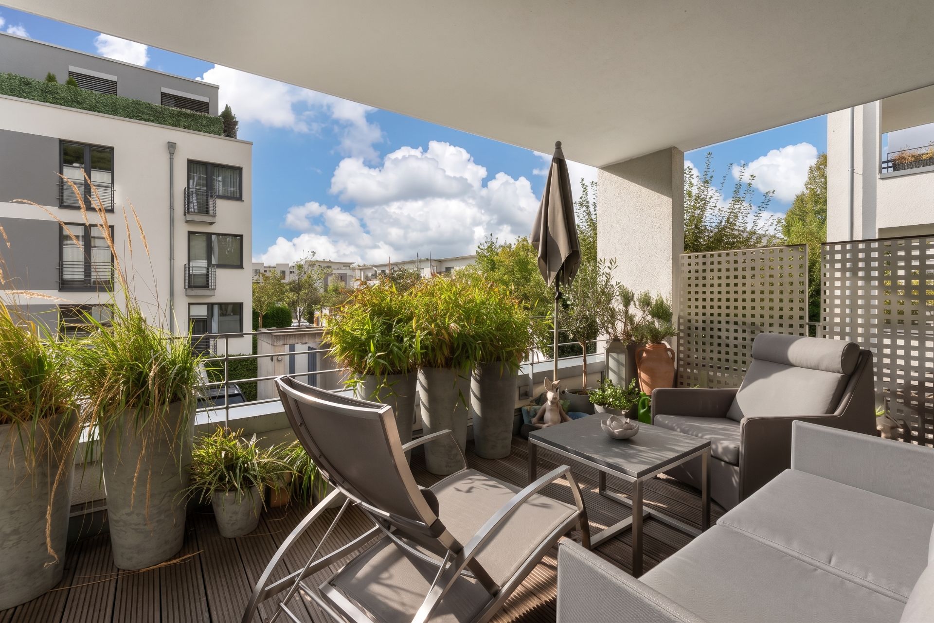 Überdachter Balkon mit Sitzmöbeln und vielen Pflanzen in großen Töpfen, Blick auf Wohngebäude.