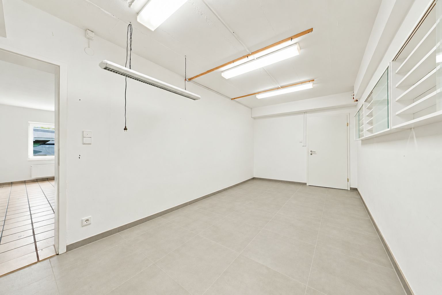 Heller leerer Raum mit Fliesenboden, weißen Wänden und Deckenlampen.