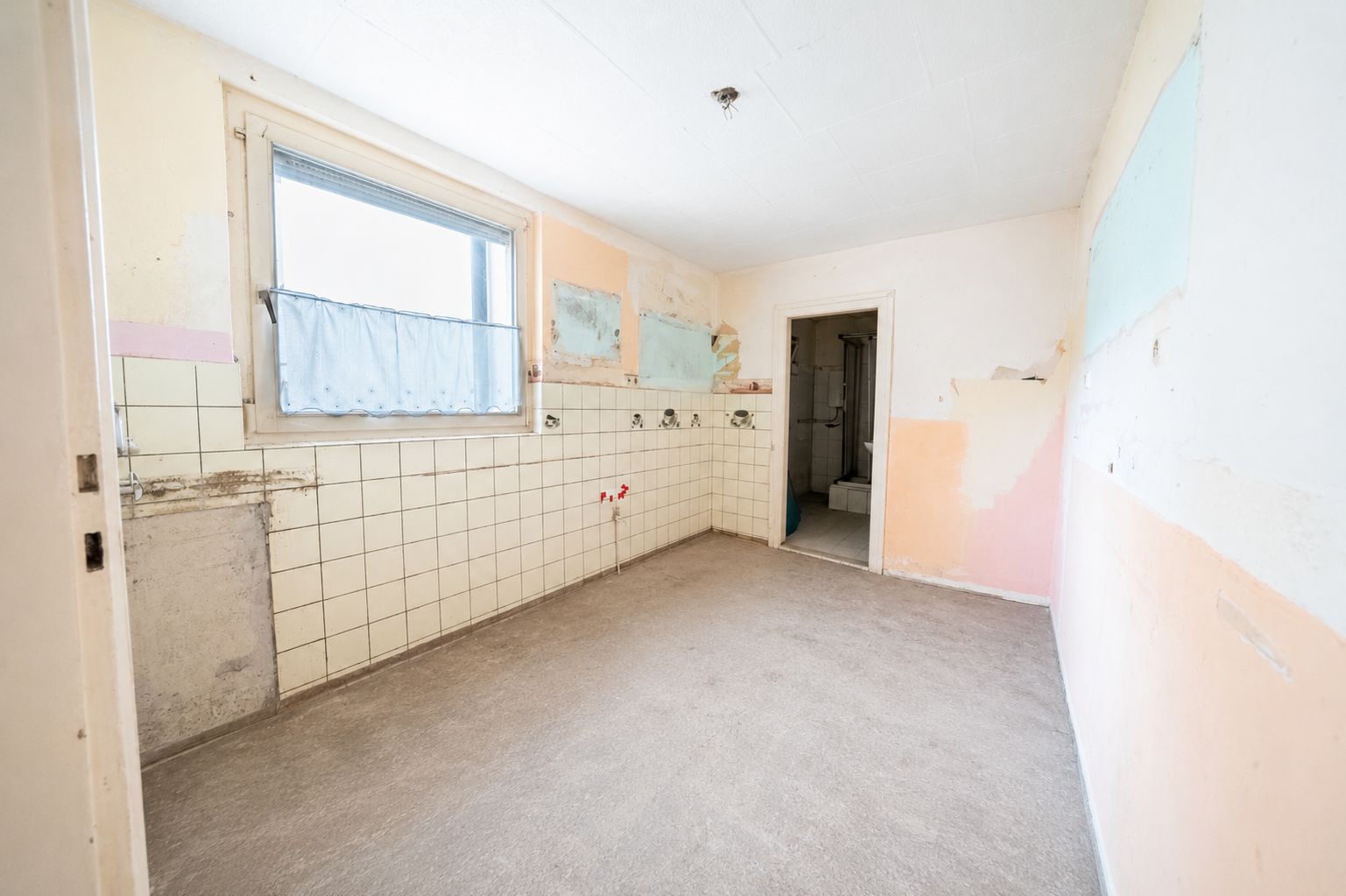 Leerer Raum mit gefliester Wand, Fenster und Tür zu einem weiteren Zimmer.