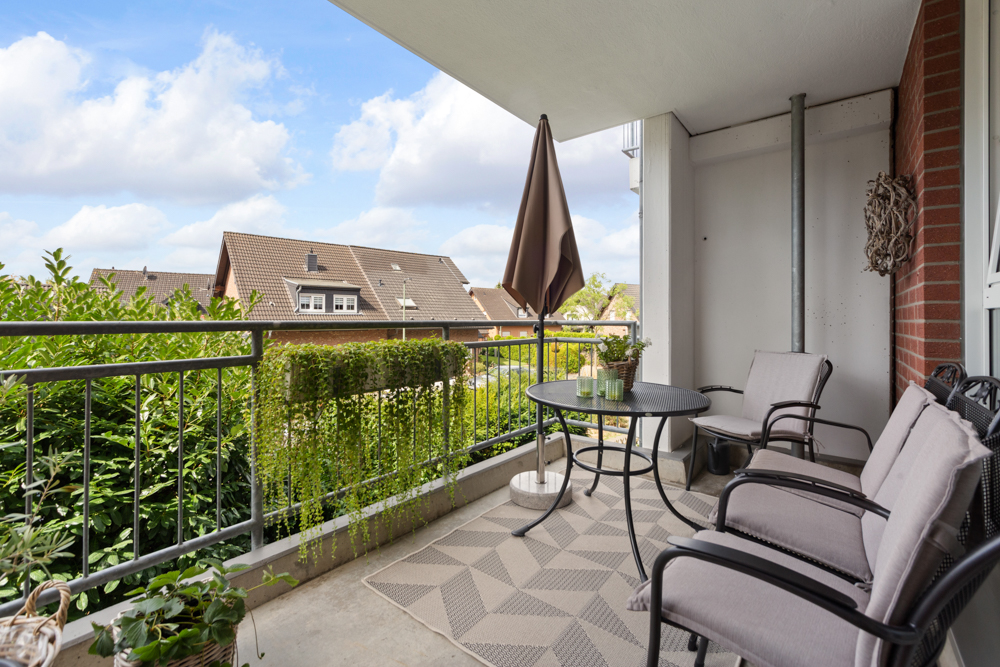 Balkon mit drei Stühlen, rundem Tisch, Teppich, Pflanzen und Blick auf Häuser und Himmel.