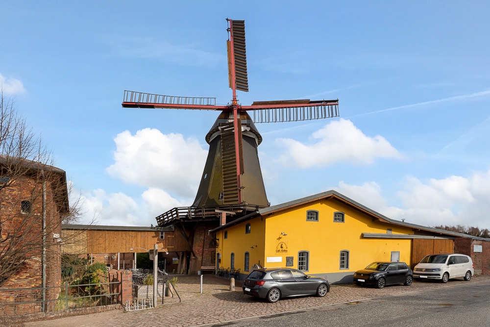 Alte Windmühle mit gelbem Nebengebäude und parkenden Autos davor bei Tageslicht.