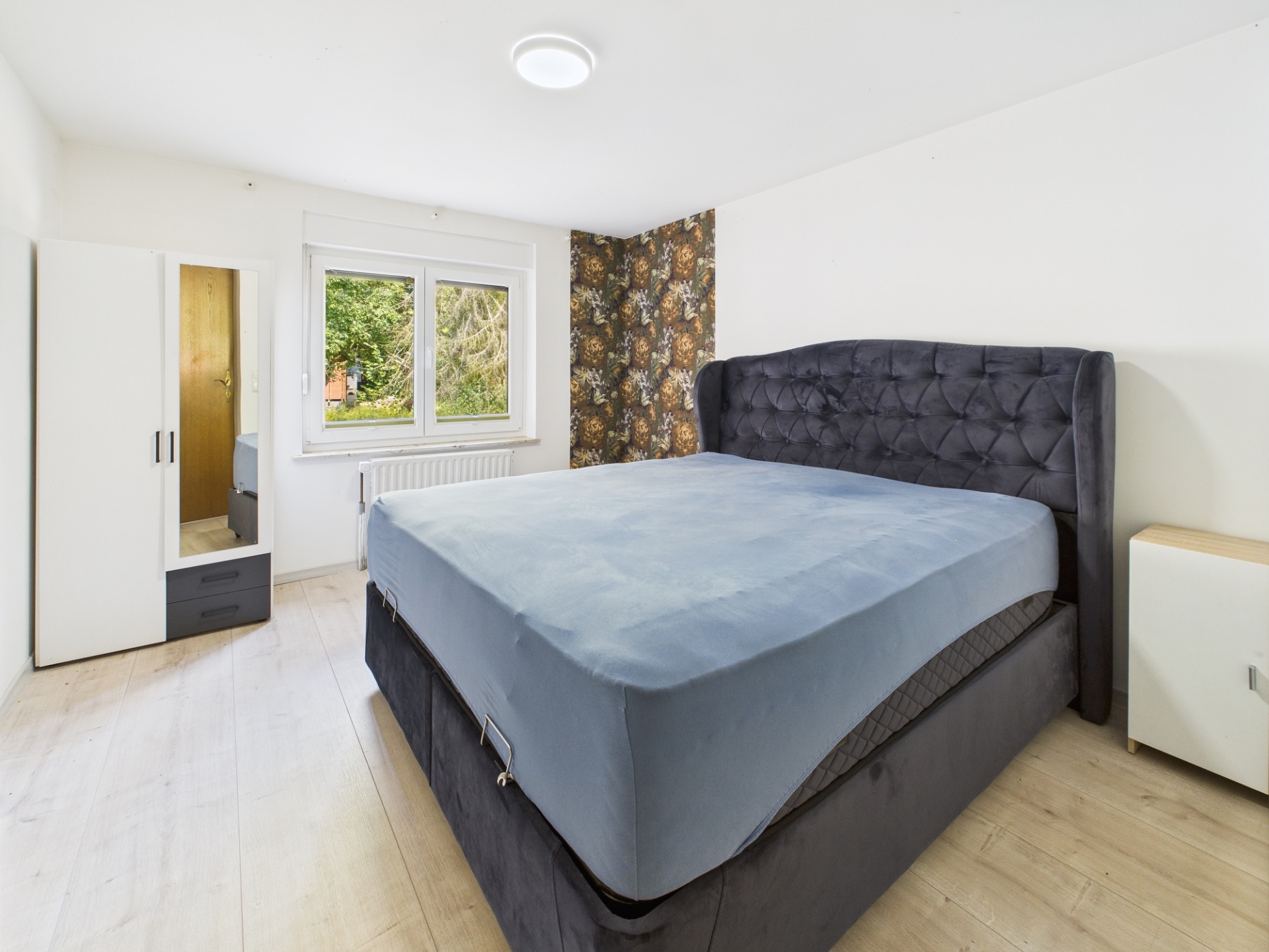 Schlafzimmer mit großem Bett, Kleiderschrank mit Spiegel und zwei Fenstern, helle Holzböden.