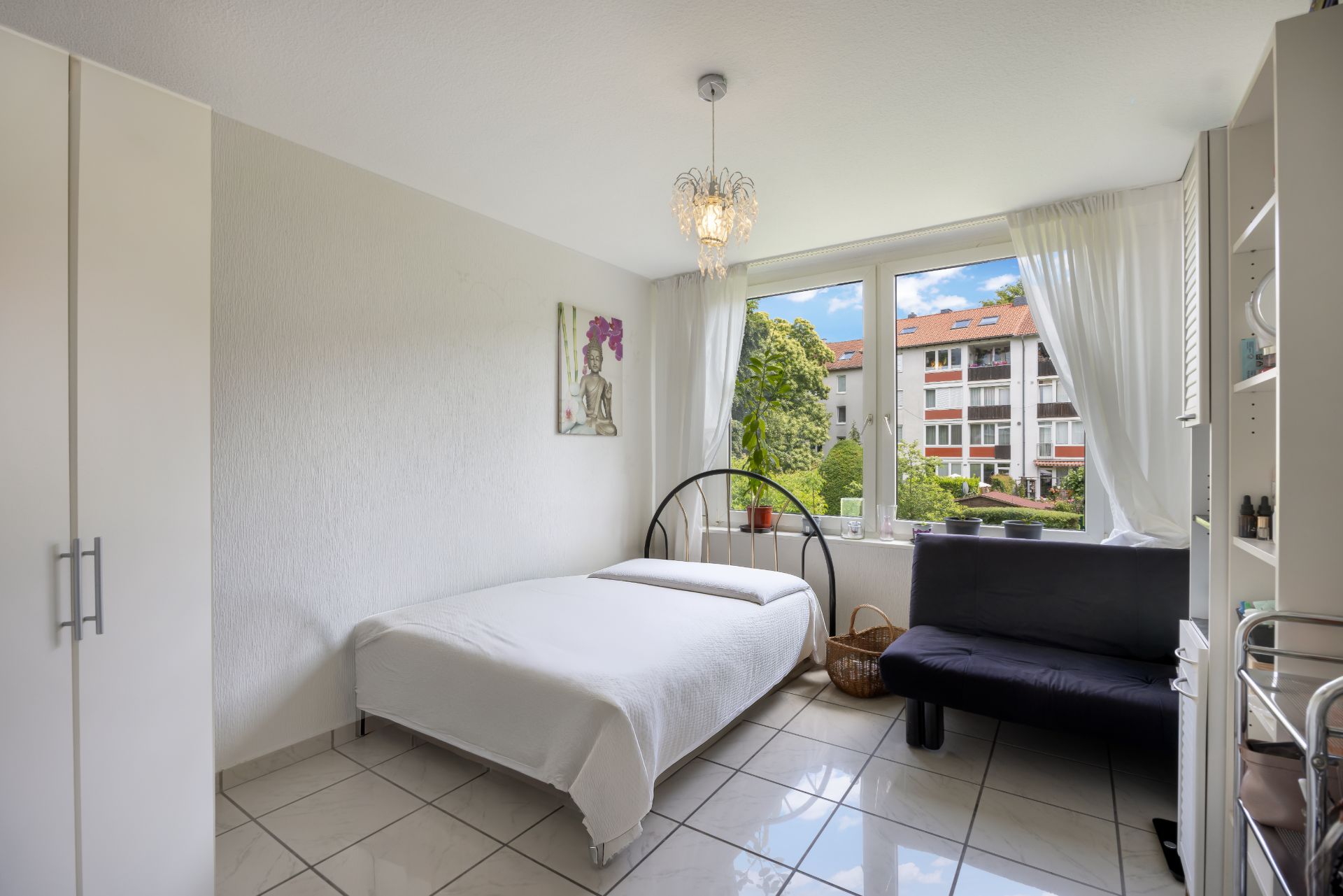 Helles Schlafzimmer mit Bett, schwarzem Sofa, großem Fenster und weißem Kleiderschrank.
