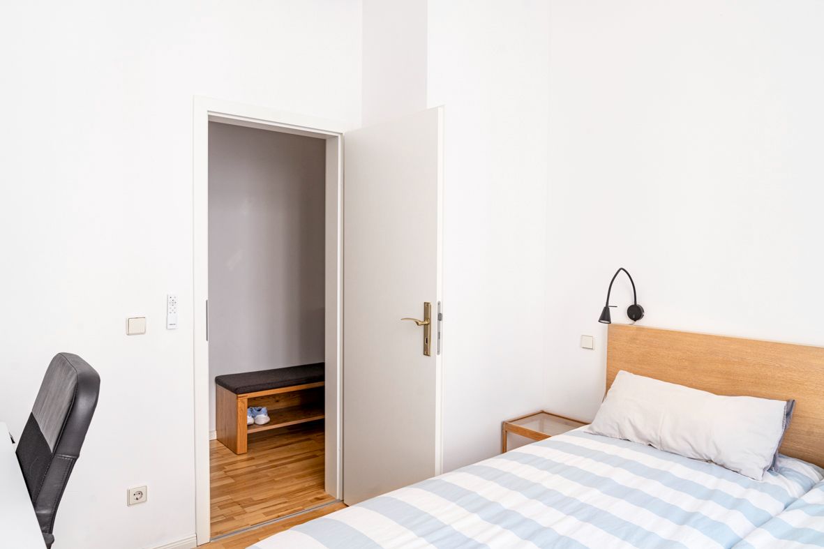 Helles Schlafzimmer mit Einzelbett, Schreibtisch, Stuhl und geöffneter Tür zu Flur mit Bank.