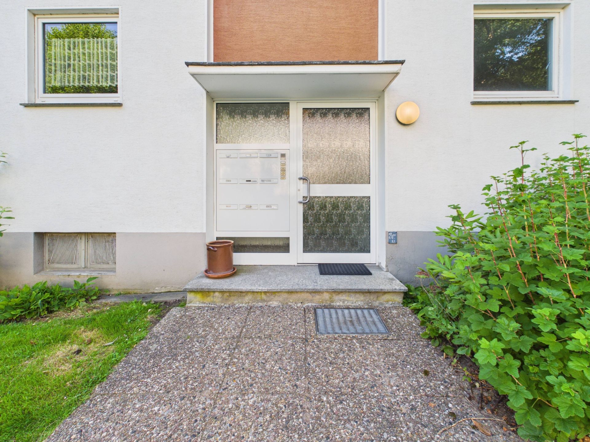 Eingangstür eines Wohnhauses mit Glastüren, Treppe und Pflanzen an den Seiten.