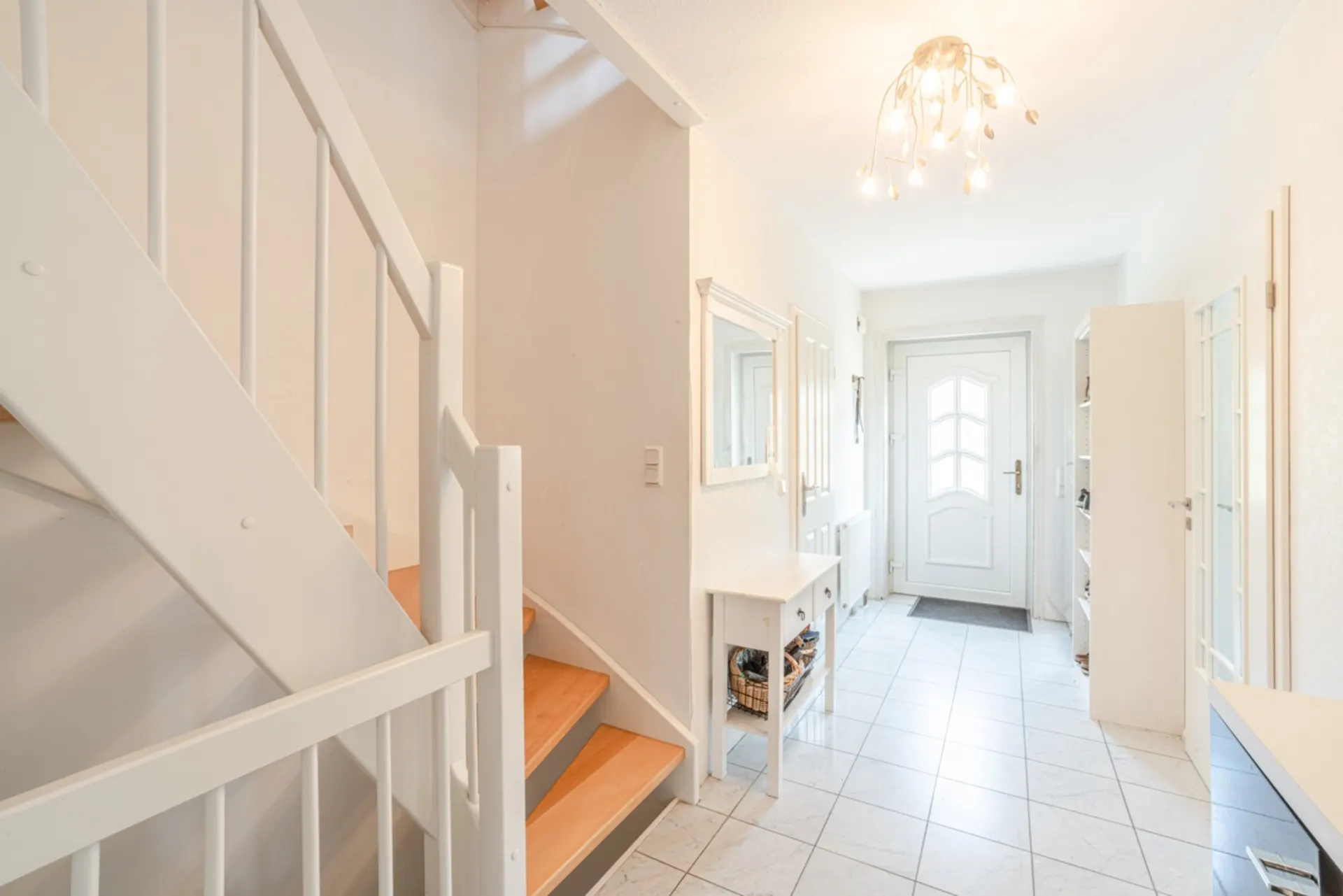 Heller Flur mit weißer Treppe, Fliesenboden, Tür, Spiegel und Tisch mit Korb.