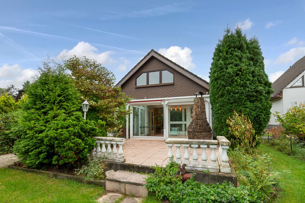 Haus mit Wintergarten und großer Terrasse, umgeben von Bäumen und Sträuchern.