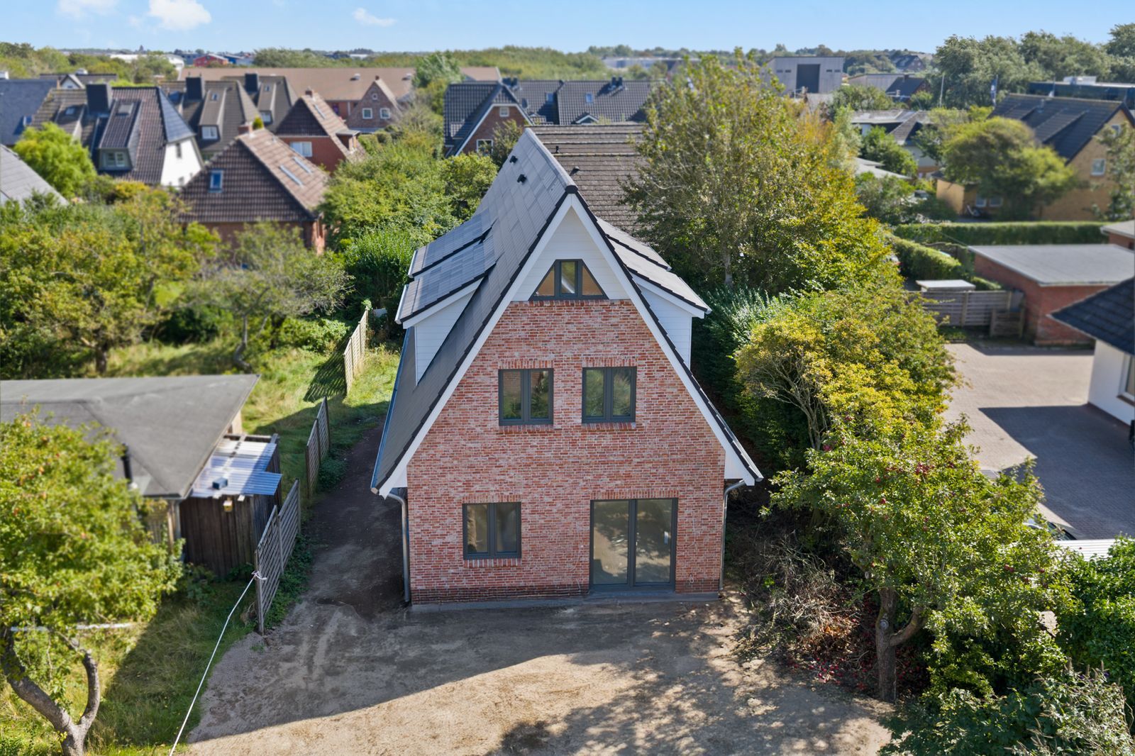 Backsteinhaus mit spitzem Dach und großen Fenstern, umgeben von Bäumen und weiteren Wohnhäusern.