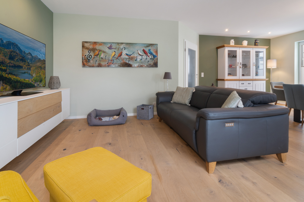Modernes Wohnzimmer mit schwarzem Sofa, gelbem Hocker, Fernseher und Bild von Vögeln an der Wand.