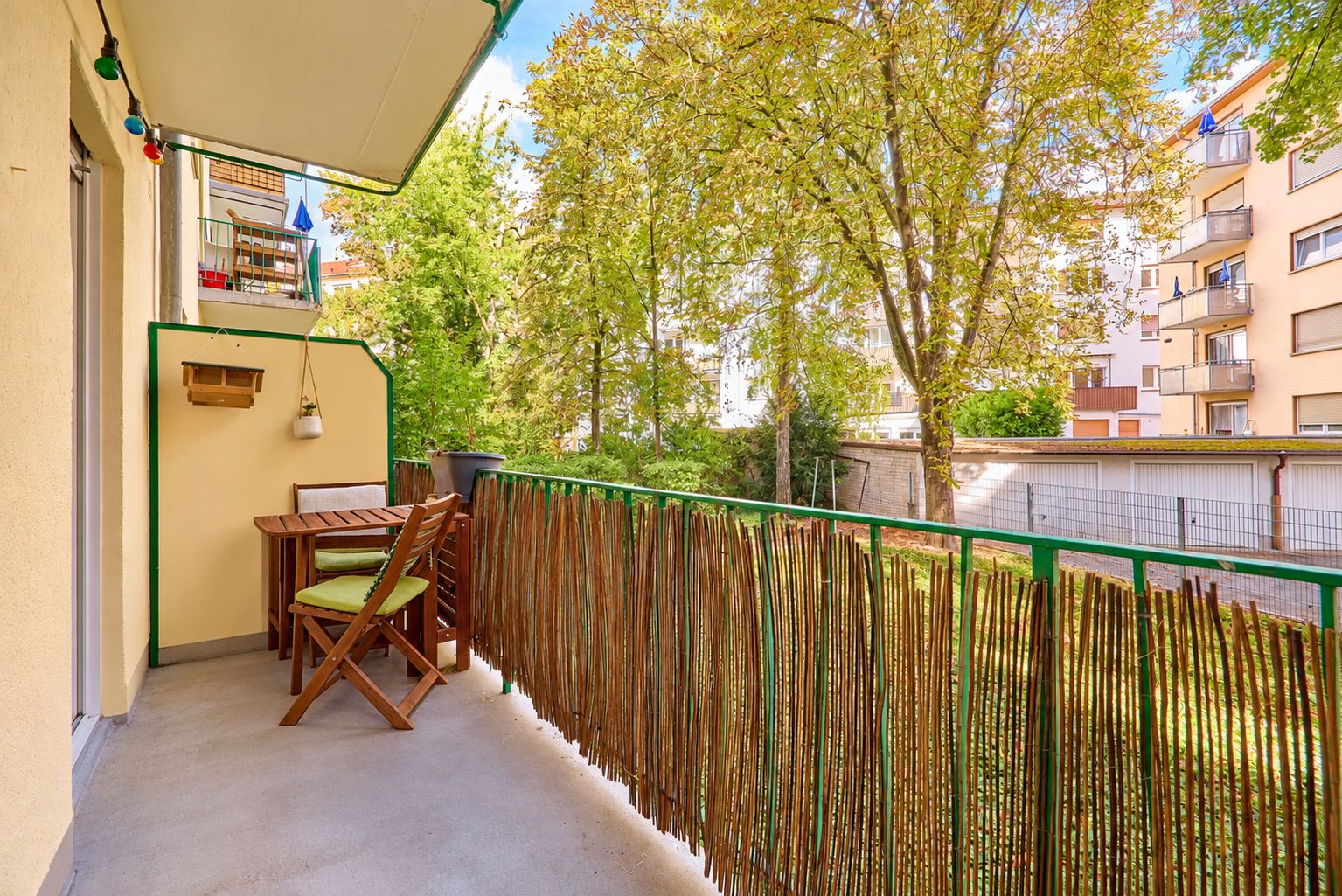 Balkon mit Holztisch und zwei Stühlen, Blick auf grüne Bäume und Wohngebäude.