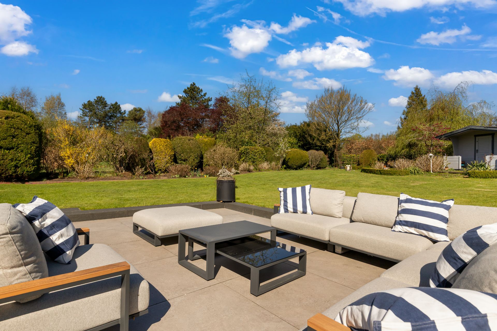 Terrasse mit modernen Sofas und Couchtisch, Blick auf gepflegten Garten bei blauem Himmel.