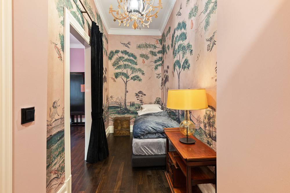 Kleines Schlafzimmer mit gemusterten Tapeten, Einzelbett und Nachttisch mit Lampe.