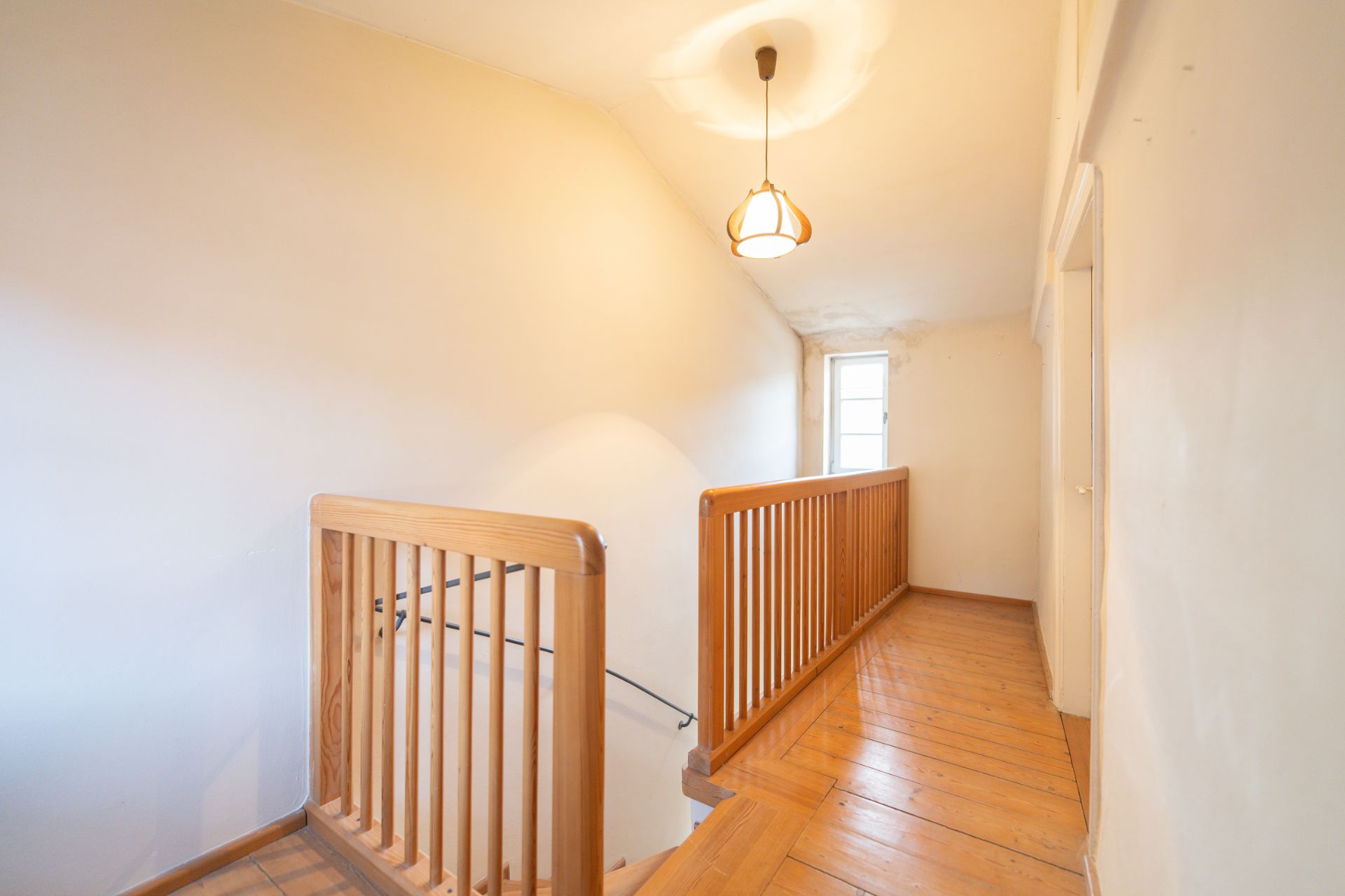 Heller Flur mit Holzgeländer, Holzfußboden und Fenster am Ende, eine Lampe an der Decke.