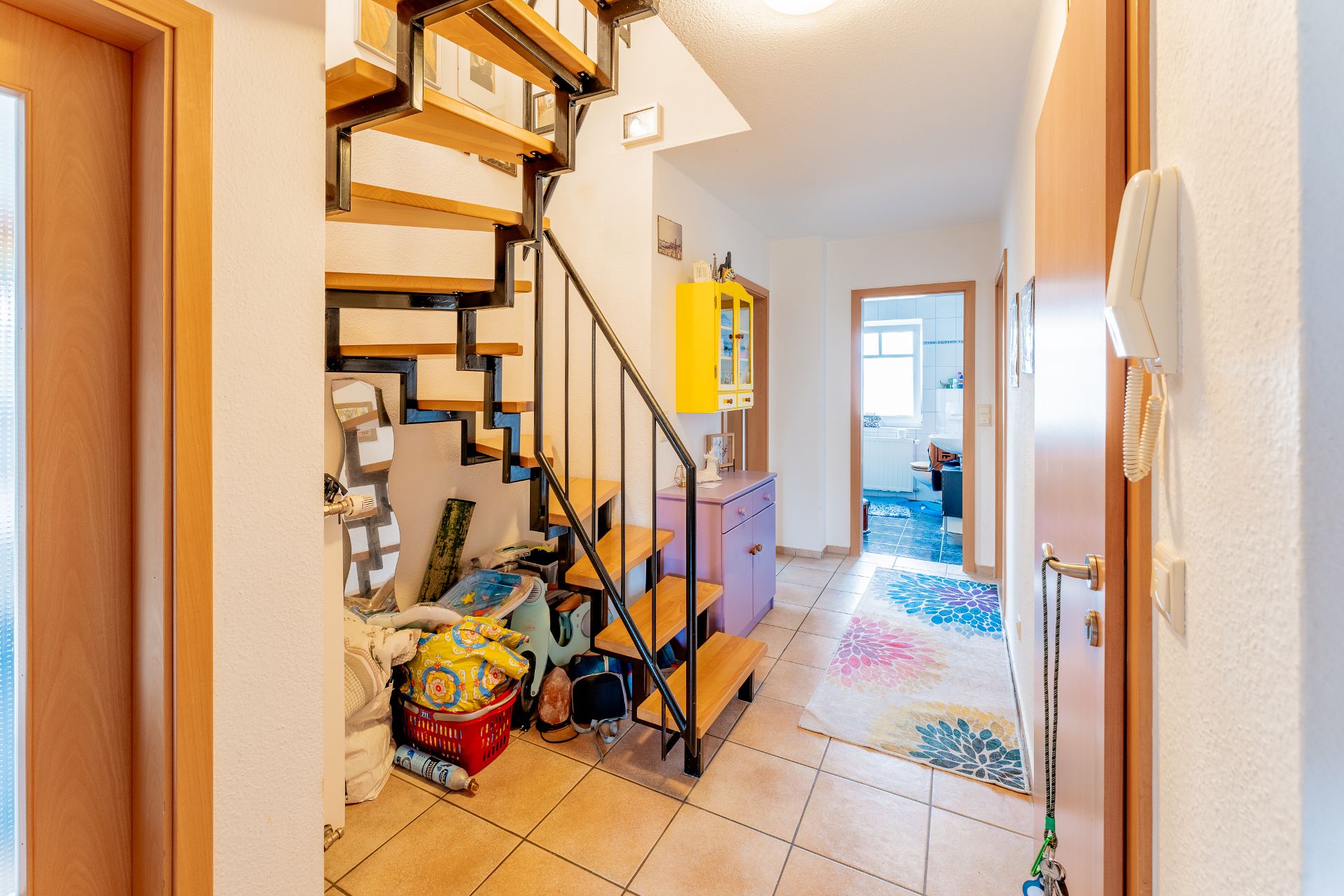 Flur mit Fliesenboden, offener Treppe, lila Kommode und farbigem Teppich.