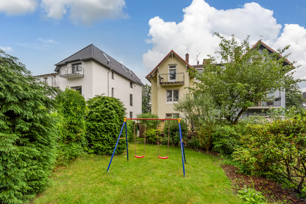 Grüner Garten mit Doppelschaukel, umgeben von Bäumen und Sträuchern, Wohnhäuser im Hintergrund.