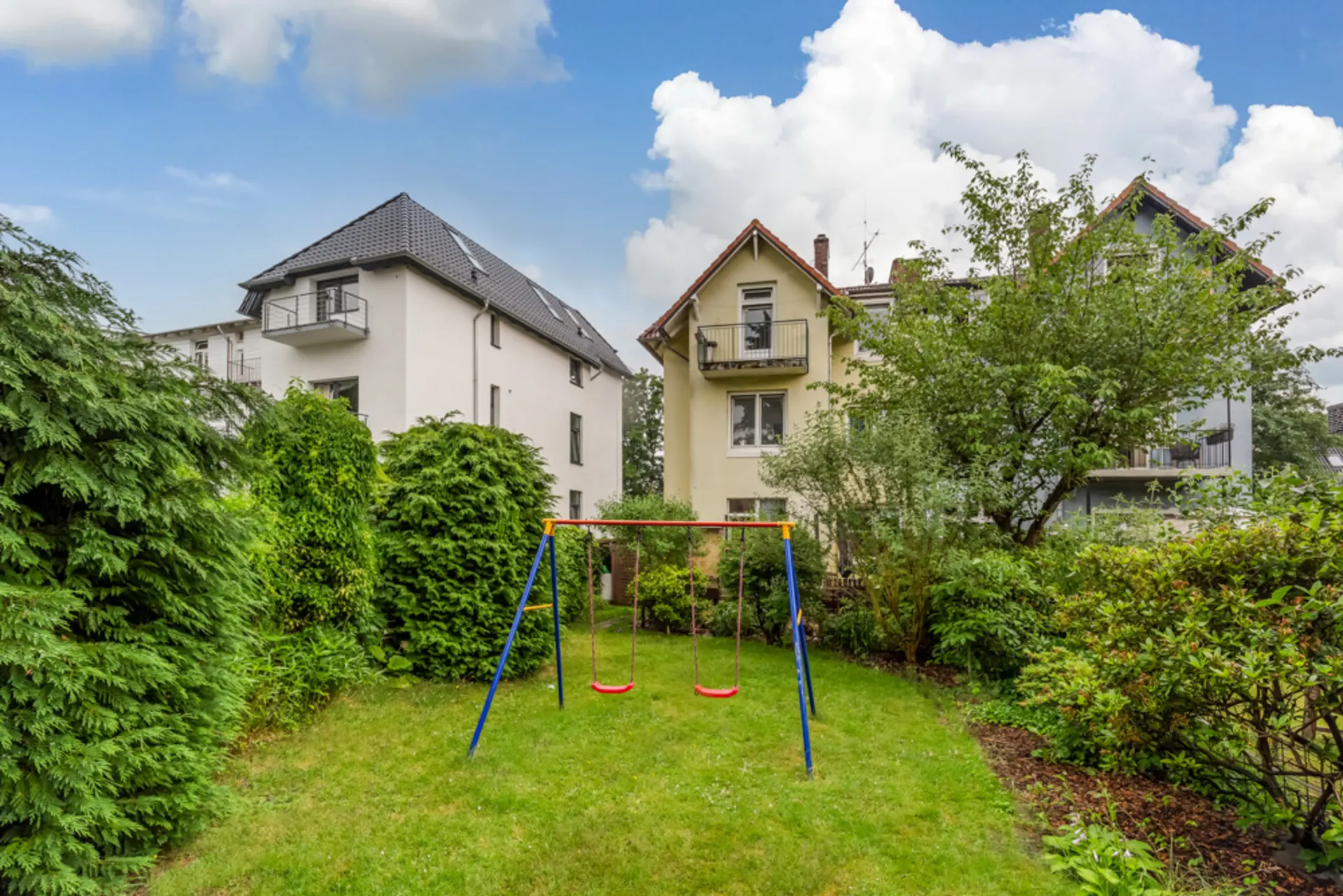Grüner Garten mit Doppelschaukel, umgeben von Bäumen und Sträuchern, Wohnhäuser im Hintergrund.