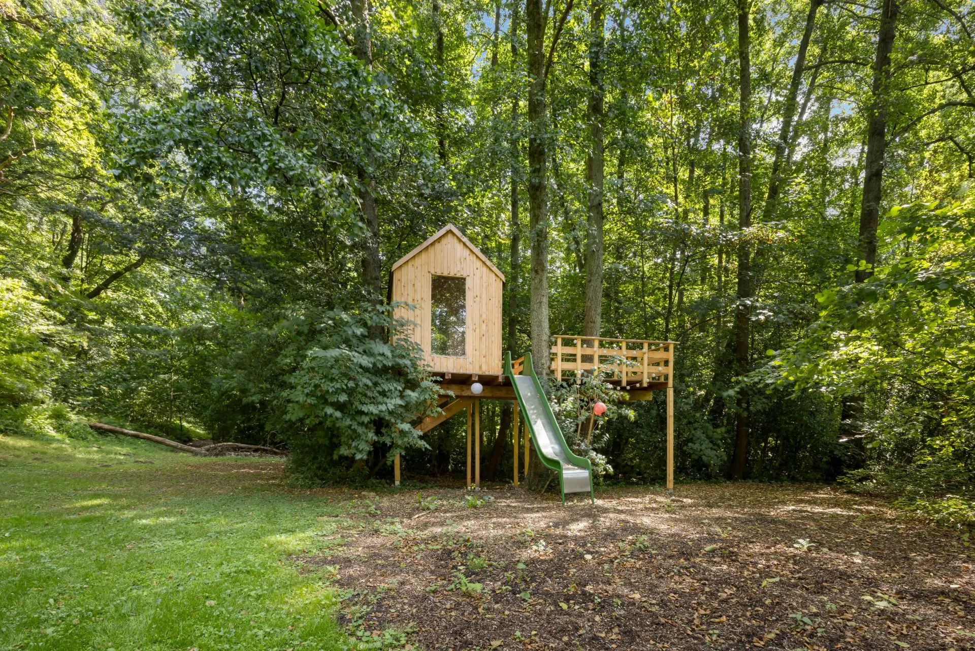Baumhaus aus Holz mit Rutsche im Garten, umgeben von hohen Bäumen und Wiese.