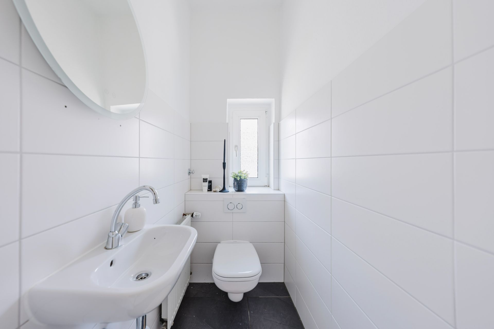 Helles, schmales Badezimmer mit Wandtoilette, Waschbecken und kleinem Fenster.
