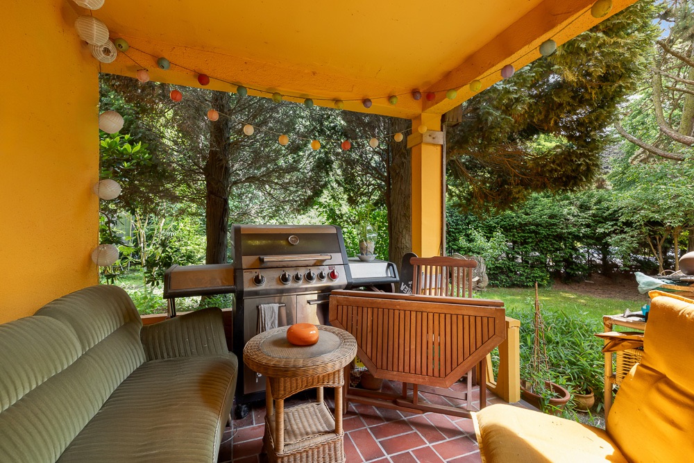 Überdachte Terrasse mit Sofa, Grill, kleinem Tisch und Gartenblick.
