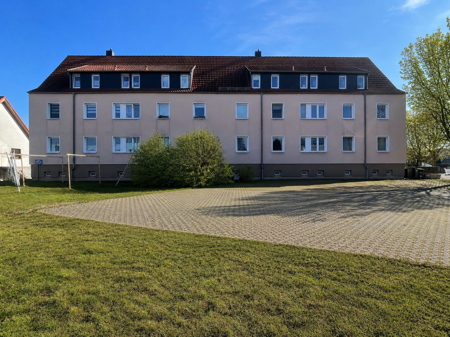 Dreistöckiges Wohnhaus mit roten Dachziegeln, davor gepflasterter Hof und Rasenfläche.