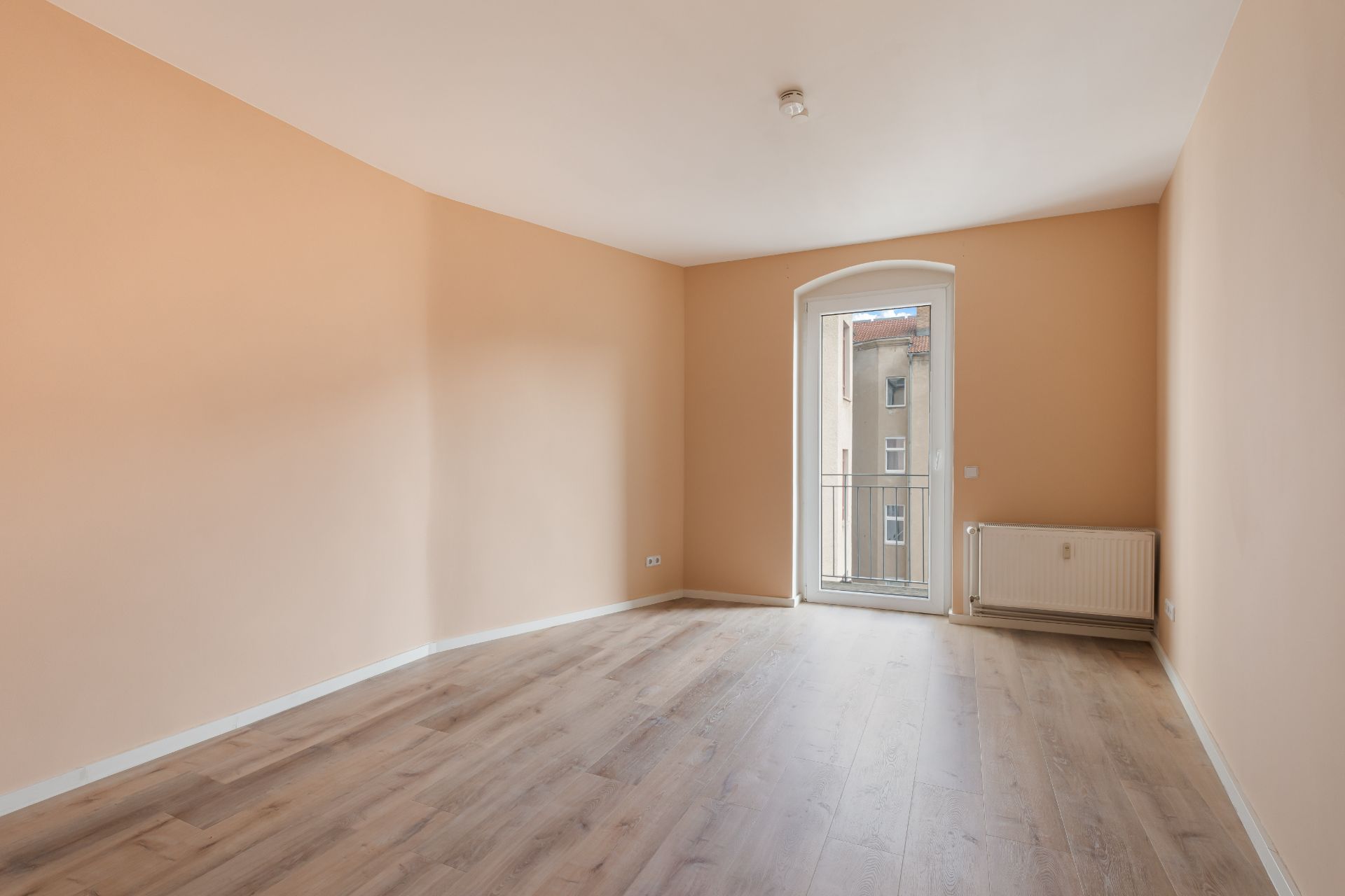 Leerer Raum mit hellen Wänden, Holzboden, Heizung und Fenstertür zum Balkon.
