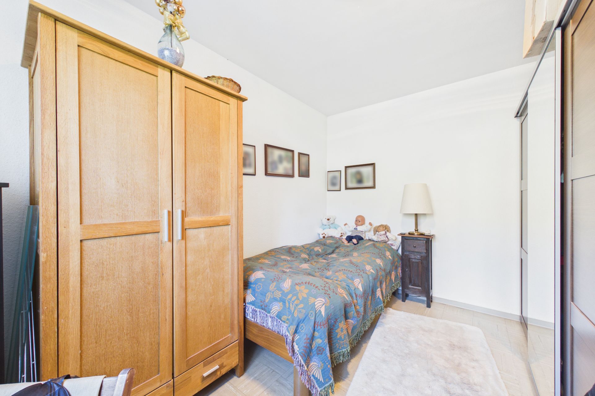 Kleines Schlafzimmer mit Holzbett, Kleiderschrank, Nachttisch, Lampe und Bildern an der Wand.