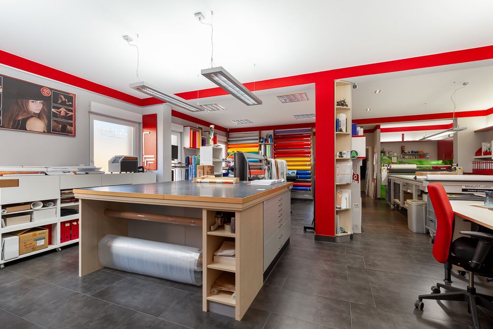 Helles Zimmer mit großem Arbeitstisch, Regalen und bunten Materialien an den Wänden.