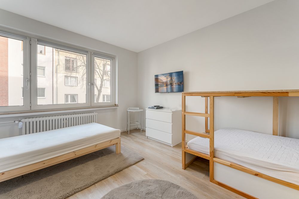 Helles Schlafzimmer mit Einzelbett, Etagenbett, Kommode und großem Fenster.